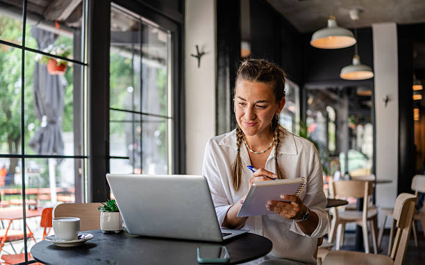 istock Young woman working on laptop in a modern cafe while taking notes and enjoying a cup of coffee on a sunny day 2224697998