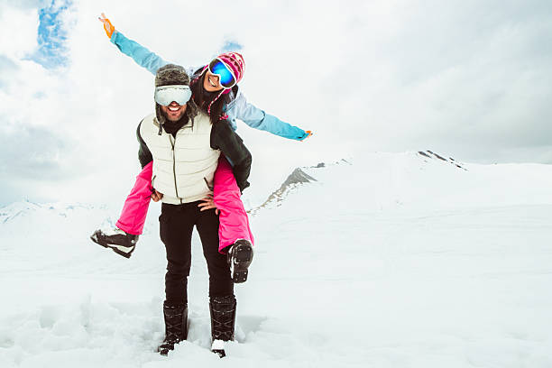 Joyful young couple walk piggyback in deep snow pose looking at camera in snowy mountain, symbolizing love, happiness, freedom, playful relationship, adventure, carefree winter escape, togetherness