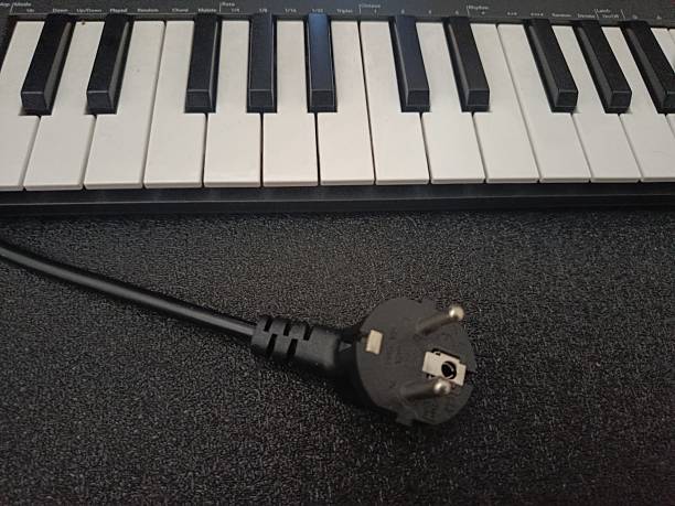 istock A close-up shot of a three-pronged black electrical plug lying on a dark surface, subtly illuminated to emphasize texture. In the background, a portion of a musical keyboard is visible, showcasing black and white keys. The juxtaposition of the power plug 2224719641