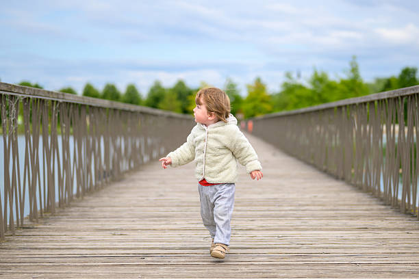 Young child in a fleece jacket walking confidently across a wooden bridge surrounded by nature.