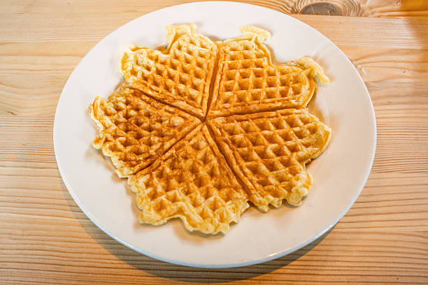 istock Fresh from a waffle iron, a toasted, golden, Norwegian waffle, on a white plate, rests on a light colored wooden countertop in a cafe at brunch 2224724275
