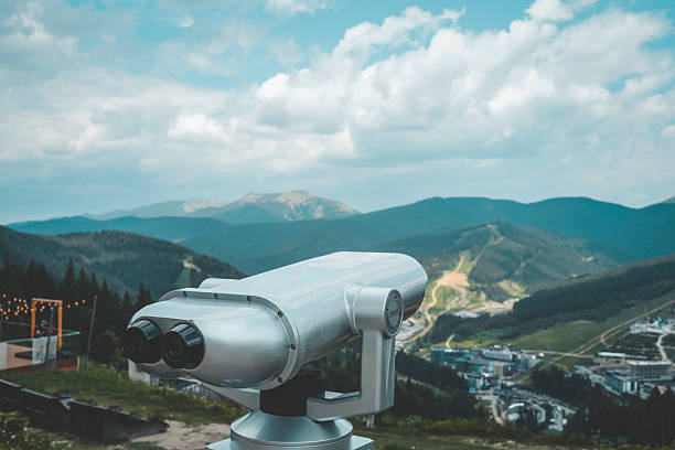 Metal binocular telescope on observation deck with scenic mountain view under cloudy sky. Tourism and sightseeing equipment at viewpoint on a clear summer day.