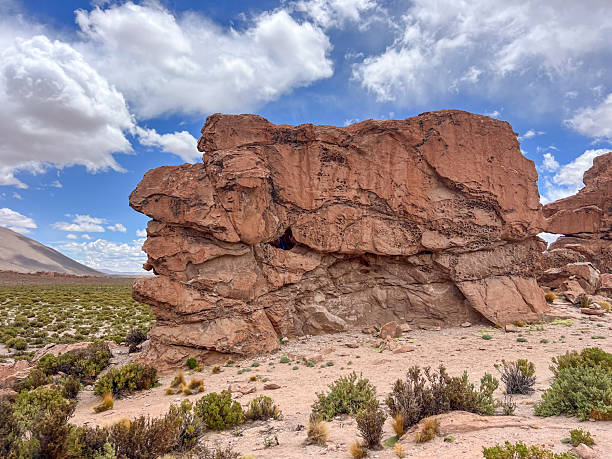 Massive red rock formation with natural erosion patterns under a partly cloudy sky. Rocky desert terrain with sparse green shrubs and distant mountains. Scenic arid landscape with geological features.