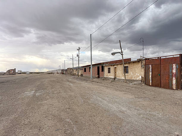 Empty dirt road lined with weathered houses and rusty gates under cloudy sky. Abandoned rural settlement with utility poles and barren landscape. Quiet desolate village scene.