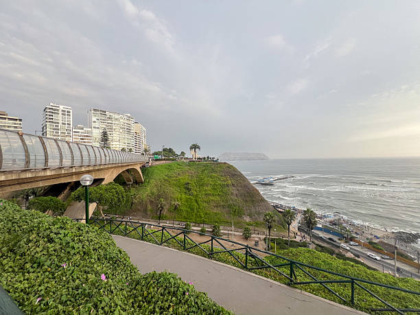 Urban coastal park with green cliffside and pedestrian bridge overlooking ocean waves. Scenic city waterfront with high-rise buildings and people walking along pathways. Outdoor nature and cityscape.