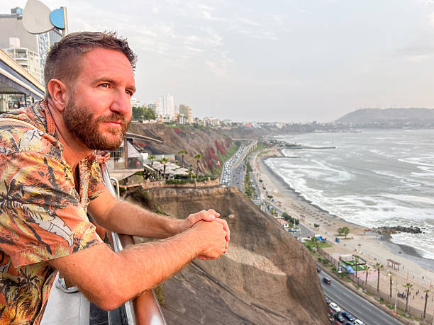 Bearded man leaning on balcony railing overlooking coastal cliffs and ocean. Relaxed adult male admiring scenic seaside cityscape with beach and road below. Person enjoying peaceful outdoor moment by the sea.