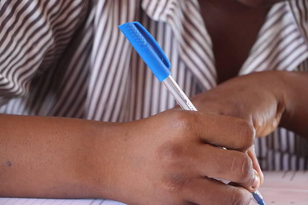 An intimate perspective on the crucial Basic Education Certificate Examination (BECE) in Ghana, featuring a close-up of a student's hand poised with a pen, actively engaged in writing. The subtle blur of classmates in the background underscores the shared, serious atmosphere of the examination. The distinct Ghanaian public or private school uniform worn by the student grounds the image firmly in its authentic West African setting. This visual encapsulates the dedication of young Ghanaians striving for educational advancement and the future generation's pursuit of knowledge in Africa.