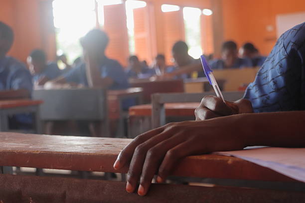 An intimate perspective on the crucial Basic Education Certificate Examination (BECE) in Ghana, featuring a close-up of a student's hand poised with a pen, actively engaged in writing. The subtle blur of classmates in the background underscores the shared, serious atmosphere of the examination. The distinct Ghanaian public or private school uniform worn by the student grounds the image firmly in its authentic West African setting. This visual encapsulates the dedication of young Ghanaians striving for educational advancement and the future generation's pursuit of knowledge in Africa.