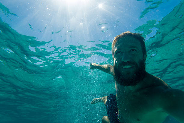 Bearded man swimming underwater in clear blue water smiling at camera. Happy young adult enjoying swimming and water activities. Person capturing underwater selfie in ocean or pool.