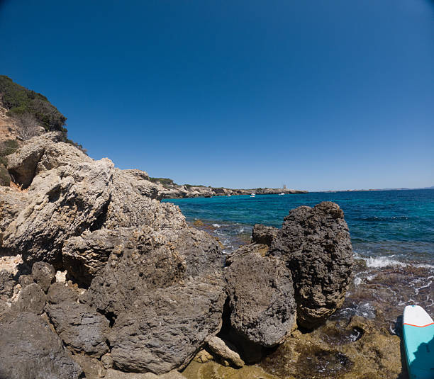 Large, rugged rocks along the shoreline with clear blue ocean waves gently crashing. Coastal landscape with rocky terrain and calm sea under a bright sunny sky. Scenic beach nature view.