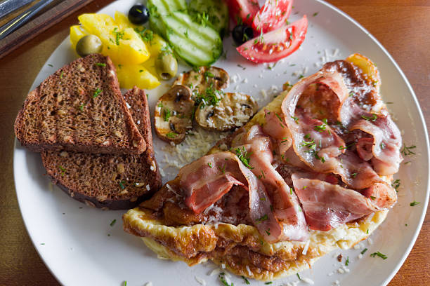 Omelet served with grilled bacon and mushrooms, olives and fresh vegetables, sprinkled with grated cheese and chopped greens, toasts of brown bread, top view in selective focus