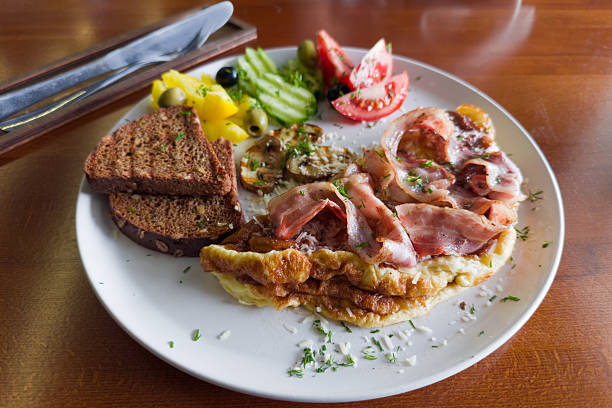 Breakfast of omelet served with grilled bacon and mushrooms, olives and fresh vegetables, sprinkled with grated cheese and chopped greens, toasts of brown bread, selective focus