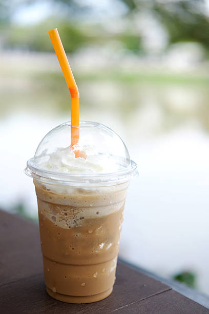 Ice latte shake coffee with whipped cream with tube in plastic glass on wooden table in natural sunlight and the river