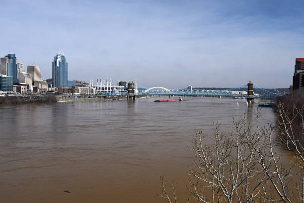 A barge traveling down the Ohio River under the Brent Spence Bridge during a flood with the Cincinnati skyline in the background
