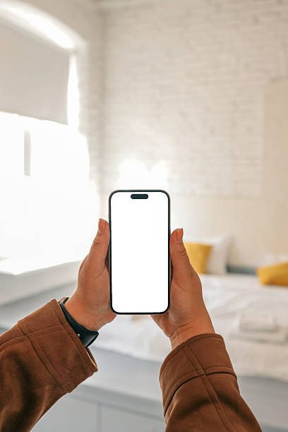 istock Hands holding smartphone with blank screen in cozy bedroom interior with white bed, yellow pillows and soft daylight. Mockup scene for mobile app or UI design 2224753680
