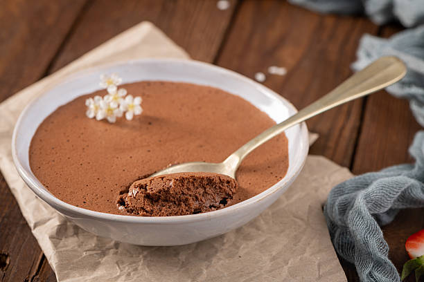 istock Elegant chocolate mousse in a shallow white dish, a golden spoon scooping a bite, adorned with tiny white blossoms, set on textured paper over a warm wooden backdrop 2224755501