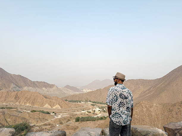 istock A man wearing a patterned shirt and hat, standing on rocks, overlooks mountainous terrain in a hazy landscape 2224778397
