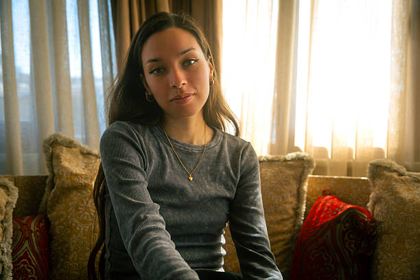A young woman sits quietly in a softly lit interior, looking into the camera with a calm and confident expression. The natural light filtering through the curtains creates a warm, intimate setting, emphasizing authenticity and emotional presence. This image is ideal for campaigns focused on mindfulness, identity, slow living, inner strength, or personal reflection.