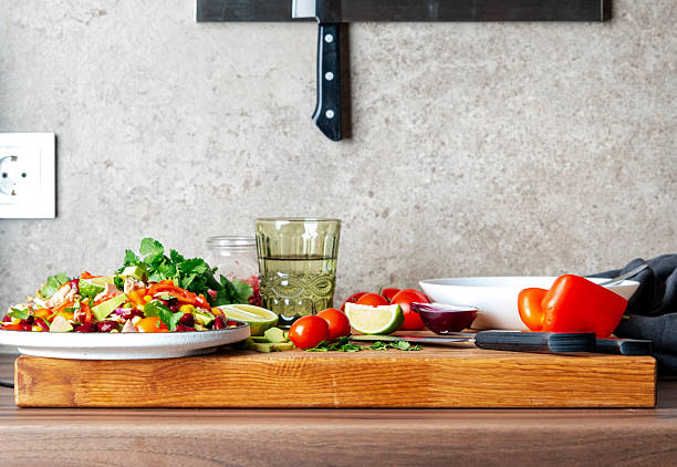 Preparing yummy salad with tuna, corn, avocado, beans, tomatoes and cilantro in plate on kitchen countertop and ingredients for preparation on wooden cutting board