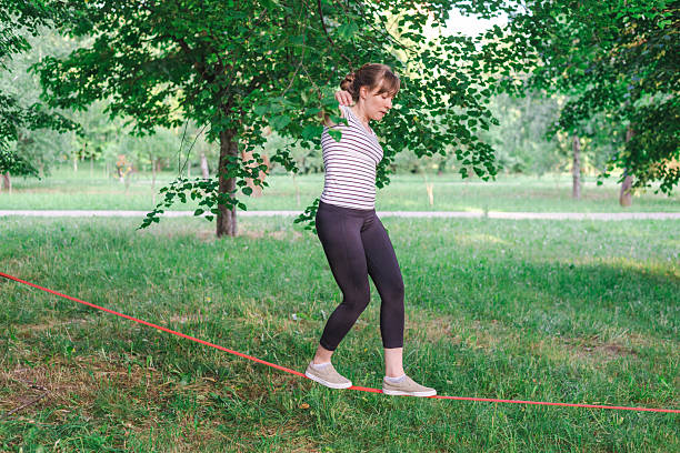 Young woman practicing slacklining in a park, balancing carefully on a red slackline tied between two trees