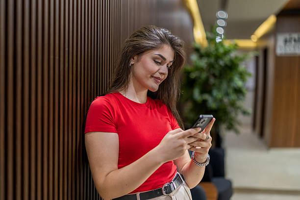 istock Young chic business woman scrolling applications on her mobile phone and resting  while standing leaning on the wood paneling wall in a corporative building. 2224791687