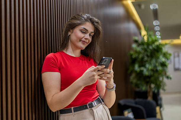 istock Young chic business woman scrolling applications on her mobile phone and resting  while standing leaning on the wood paneling wall in a corporative building. 2224791700
