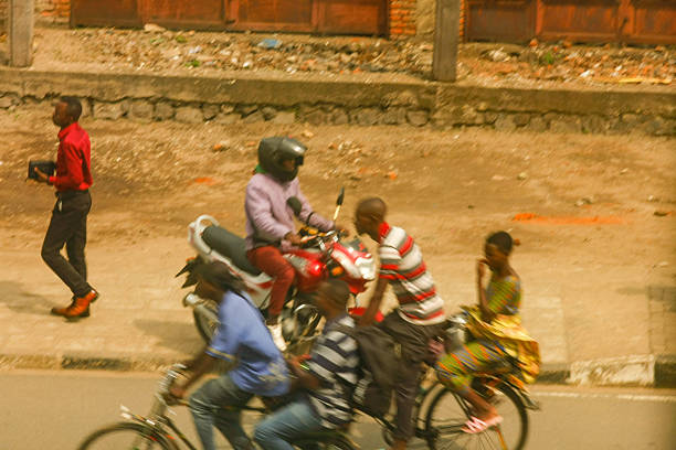 A dynamic and authentic snapshot of daily life on a bustling street in Rubavu, Rwanda. The image captures the energy and movement of the city, featuring locals navigating the thoroughfare on motorcycles and bicycles, alongside a pedestrian. The warm, natural light illuminates the vibrant atmosphere and provides a glimpse into local transportation and daily routines in this Rwandan city.