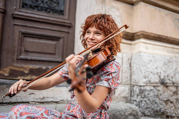 A cheerful young woman with curly red hair plays the violin outside a historic building, showcasing musical talent and joyful expression while embracing an artistic and vintage ambiance.
