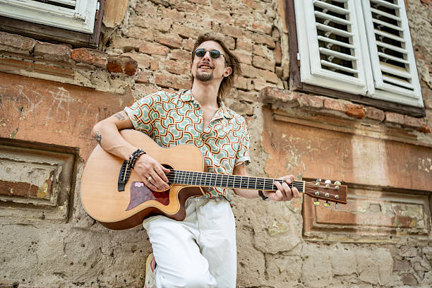 A stylish musician enjoys strumming his acoustic guitar outdoors near a rustic brick wall, exuding a laid-back and creative vibe in a vintage-inspired shirt, expressing his artistic and carefree personality.