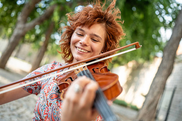 A cheerful young woman with curly auburn hair playing the violin under the shade of lush green trees on a sunny day, showcasing passion and love for music amidst a serene outdoor environment.
