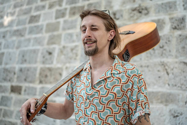 A cheerful guitarist with light brown hair winks while carrying an acoustic guitar, locking attention with a stylish vintage patterned shirt. The stone wall background complements the lively and retro atmosphere.