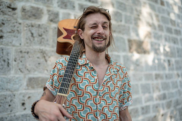 A cheerful man in patterned clothing holds a guitar, standing by a stone wall. He exudes a positive and relaxed vibe, perfectly framed in an outdoor natural light setting.