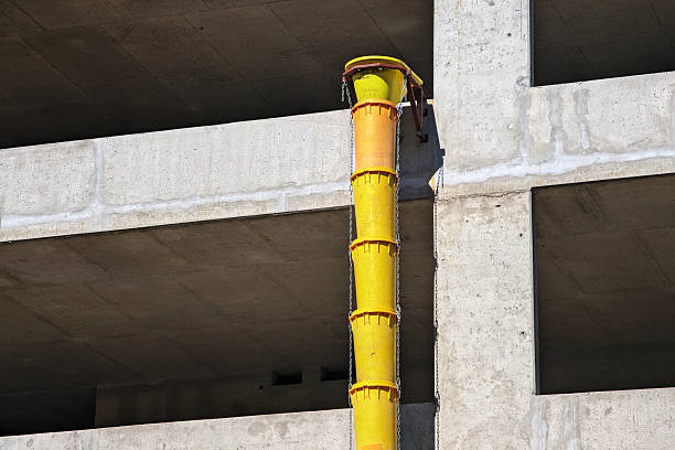 A bright yellow debris chute with metal reinforcement bands extends down the side of a concrete building under construction, allowing workers to safely dispose of rubble from upper floors. This essential construction equipment represents the careful engineering behind urban development, where safety and efficiency work together to transform raw structures into finished buildings.