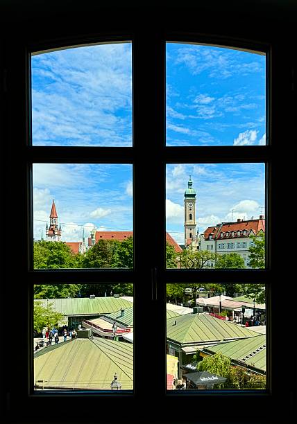 A picturesque view of Munich’s historic city center seen through a window. The scene captures rooftops of Viktualienmarkt and iconic towers against a vivid blue sky, perfectly framed by the black window grid.