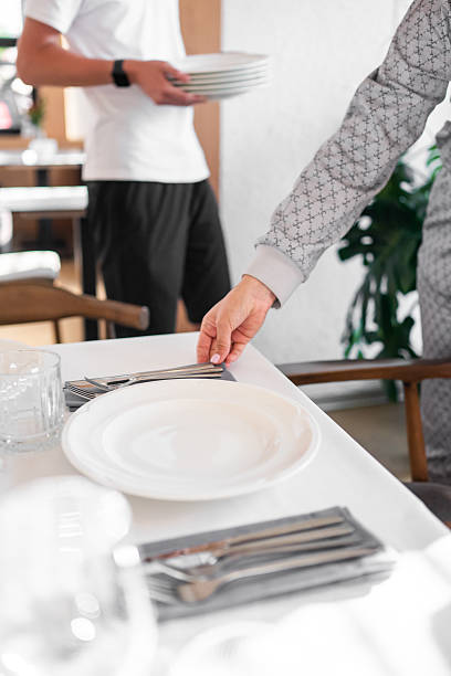 istock Restaurant staff preparing table for meal service, placing cutlery and plates, creating a clean and organized dining setup in a bright indoor setting. 2224811700