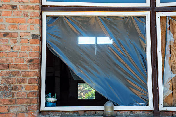 A weathered window with a plastic cover flaps slightly in the breeze, revealing a hint of greenery outside from an old brick building