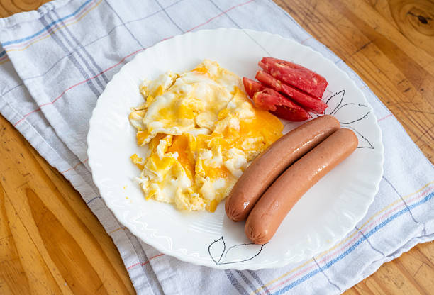Traditional breakfast plate with scrambled eggs, sausages, fresh tomato slices, served on a white plate with a homey kitchen background. High quality photo