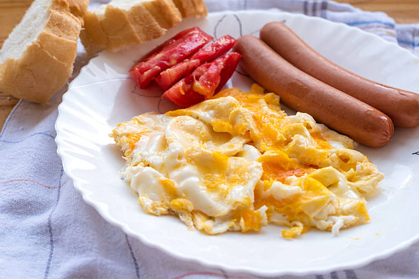 Traditional breakfast plate with scrambled eggs, sausages, fresh tomato slices, and bread, served on a white plate with a homey kitchen background. High quality photo