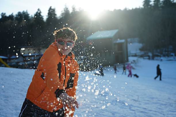 Teen in orange jacket joyfully throwing snow in a winter landscape. The image captures a playful moment with sunlight and forest in the background, highlighting fun in the snow.