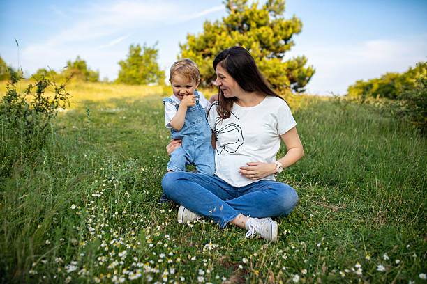 A young pregnant woman sits on the grass in a vibrant spring meadow surrounded by wildflowers and lush greenery. She smiles warmly as she embraces her cheerful little boy, who stands playfully beside her wearing a white T-shirt and adorable denim overalls. The moment captures the carefree joy of childhood, the deep bond between mother and son, and the beauty of spending time together in nature.