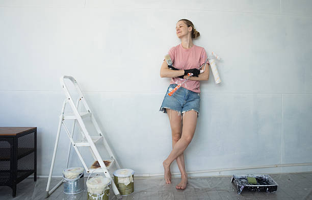 istock Young woman painting indoors wall with roller using stepladder 2224857794