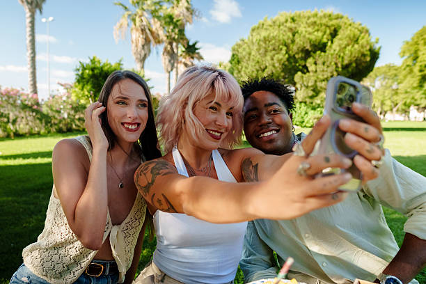 Three cheerful multi-ethnic friends are taking a selfie with a smartphone during a picnic in a park