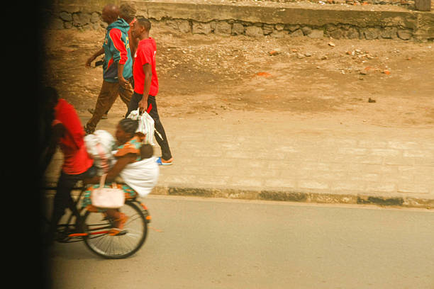 This vibrant street scene in Rubavu, Rwanda, features several people engaged in daily activities. On the left, a partially visible person is on a bicycle. In the mid-ground, two men walk, one in a red t-shirt carrying a patterned bag, and another in a blue and red hooded jacket. On the right, a person on a bicycle carries another individual wrapped in fabric. The background shows a brick building with wooden doors and a dusty, unpaved area, typical of an urban setting. The image captures the dynamic energy of urban life and various modes of transportation in Rubavu.