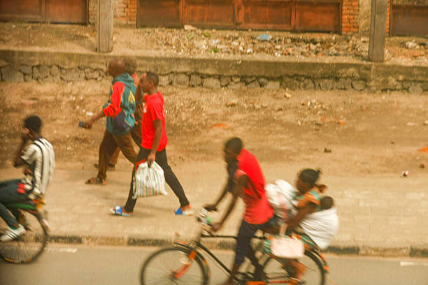 This vibrant scene in Rubavu, Rwanda, features several people engaged in daily activities. On the left, a person on a bicycle with a striped shirt is partially visible. In the mid-ground, two men walk, one in a red t-shirt holding a patterned bag and another in a blue and red hooded jacket. On the right, a person on a bicycle carries another individual wrapped in fabric. The background shows a brick building with wooden doors and a dusty, unpaved area. The image captures the dynamic energy of urban life and various modes of transportation in Rubavu.