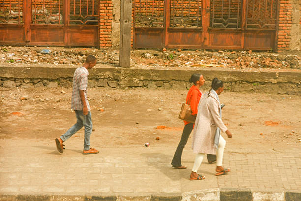 Three individuals walk along a dusty area beside a paved sidewalk in Rubavu, Rwanda. On the left, a man in a plaid or patterned shirt and jeans walks towards the right. In the center, a woman in an orange top and dark pants with a brown bag walks. On the right, a woman in a light-colored long coat and lighter pants walks, possibly holding a phone. The background features a brick building with wooden doors and an unpaved, debris-strewn area, characteristic of an urban setting. The image captures a candid moment of everyday life and movement in the city.