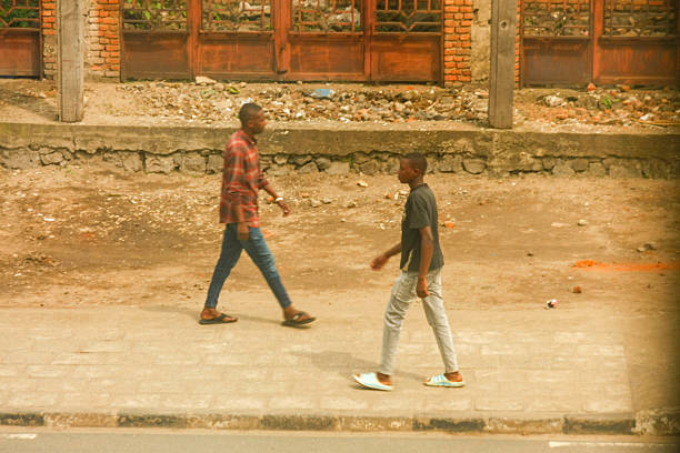 Two men walk along a paved sidewalk in Rubavu, Rwanda. The man on the left wears a red and black plaid shirt and blue jeans, while the man on the right wears a dark grey t-shirt and light grey pants. The background features a brick building with prominent wooden doors and an unpaved, dusty area, characteristic of an urban setting. The image captures a candid moment of everyday life and movement in the city.