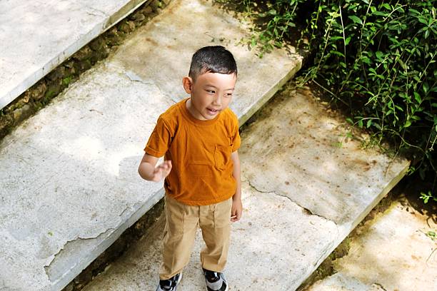 A little boy in orange shirt and beige pants looks away while standing on stairs outside, expressing calm thoughtfulness.