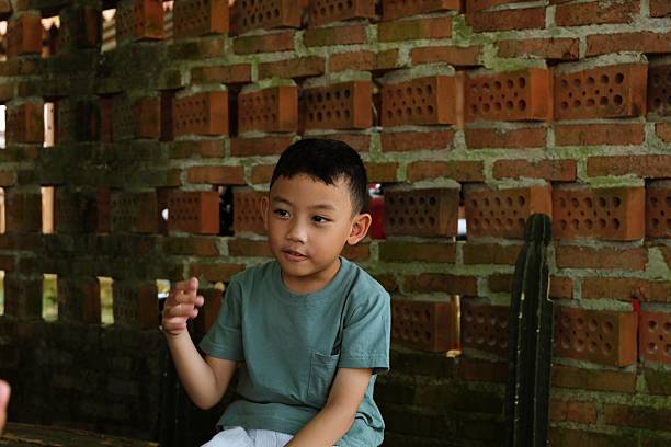 A cheerful boy gestures with his hand while sitting on a bench against a rustic brick wall, enjoying a casual moment.