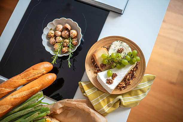 A kitchen counter displaying French culinary staples including cheese, grapes, walnuts, fresh baguettes, and snails on a ceramic plate. The vibrant arrangement evokes a sense of traditional French cooking.