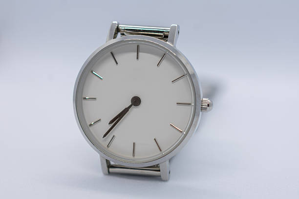 A stylish wristwatch features a simple white face and mesh band, hanging beside a window adorned with green plants, capturing a serene atmosphere filled with natural light.